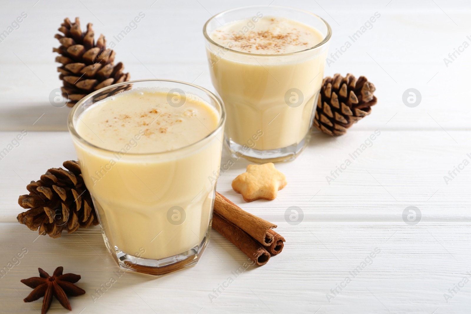 Photo of Tasty Christmas cocktail (eggnog) in glasses, pine cones and spices on white wooden table, closeup. Space for text