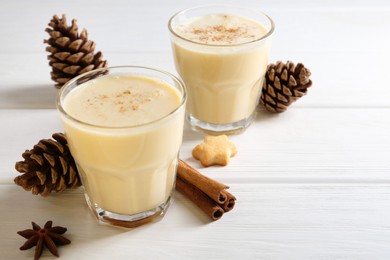 Photo of Tasty Christmas cocktail (eggnog) in glasses, pine cones and spices on white wooden table, closeup. Space for text