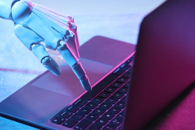 Photo of Artificial intelligence. Robot hand using laptop on table in neon lights, closeup