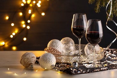 Photo of Glasses with red wine and beautiful Christmas decor on white table against blurred lights, space for text