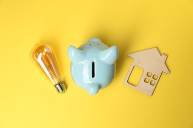 Photo of Piggy bank, light bulb and house model on yellow background, flat lay. Energy saving concept