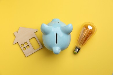 Photo of Piggy bank, light bulb and house model on yellow background, flat lay. Energy saving concept