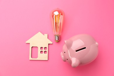 Photo of Piggy bank, light bulb and wooden house model on pink background, flat lay. Energy saving concept