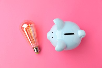 Photo of Piggy bank and light bulb on pink background, flat lay. Energy saving concept