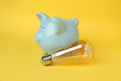 Photo of Piggy bank and light bulb on yellow background, closeup. Energy saving concept