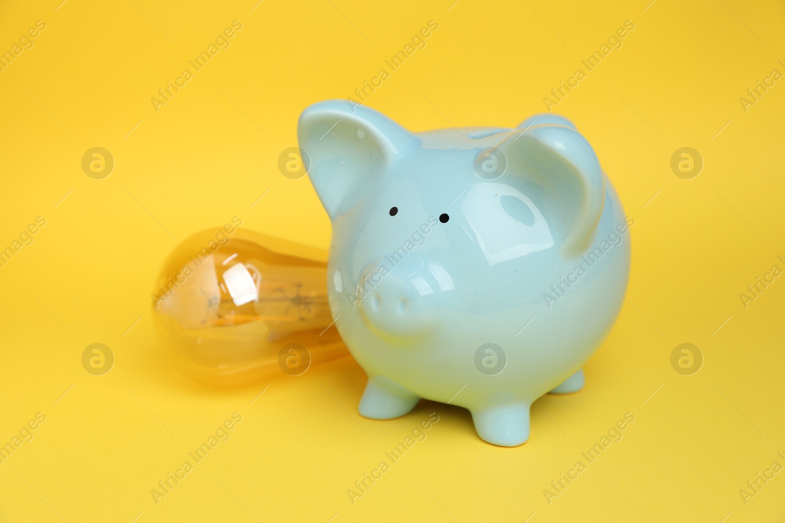 Photo of Piggy bank and light bulb on yellow background, closeup. Energy saving concept