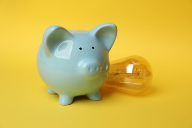 Photo of Piggy bank and light bulb on yellow background, closeup. Energy saving concept