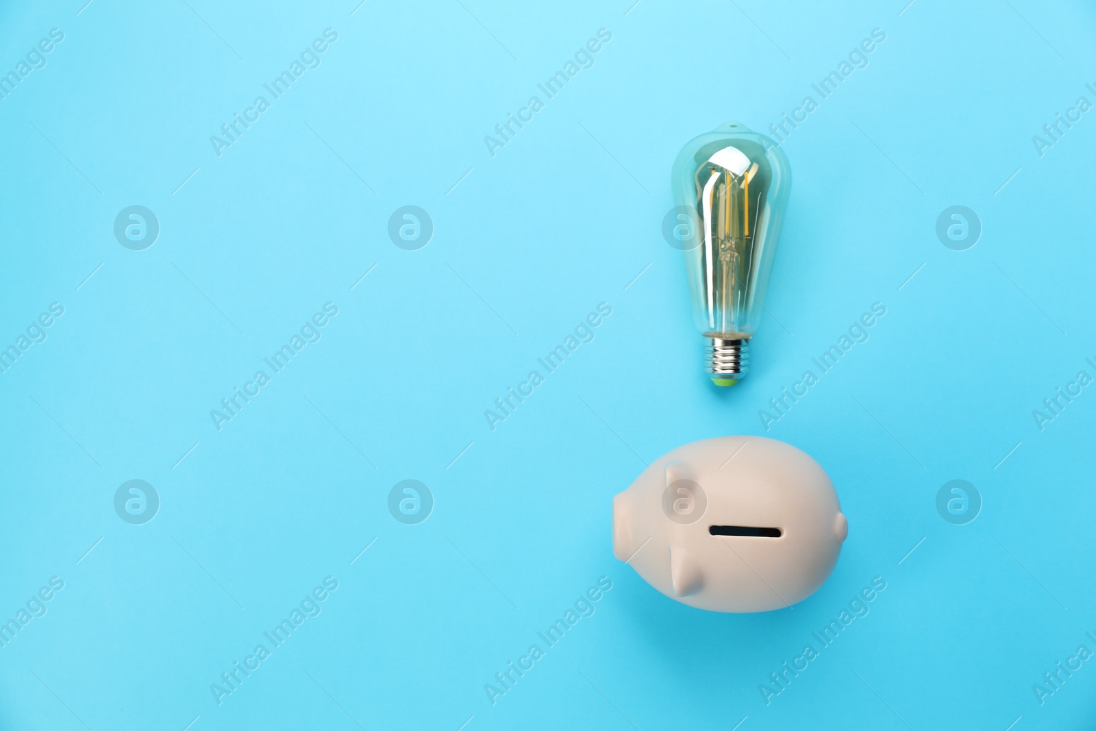 Photo of Piggy bank and lightbulb on light blue background, flat lay with space for text. Energy saving concept