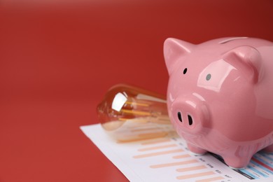 Photo of Energy saving concept. Piggy bank, light bulb and charts on red background, closeup. Space for text