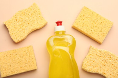 Many sponges and bottle of detergent on beige background, flat lay