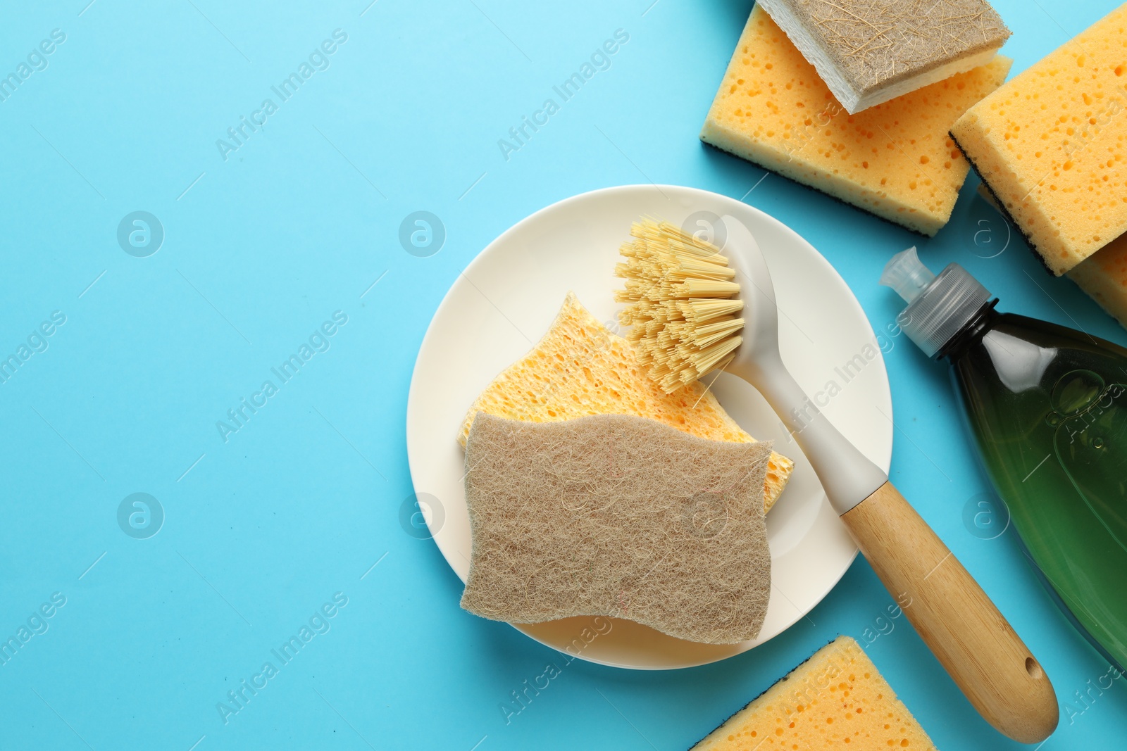 Photo of Sponges, plate, detergent and brush on light blue background, flat lay. Space for text