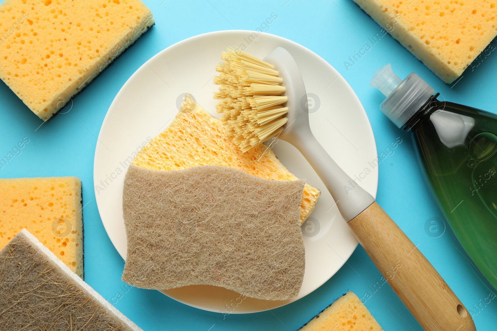 Photo of Sponges, plate, detergent and brush on light blue background, flat lay