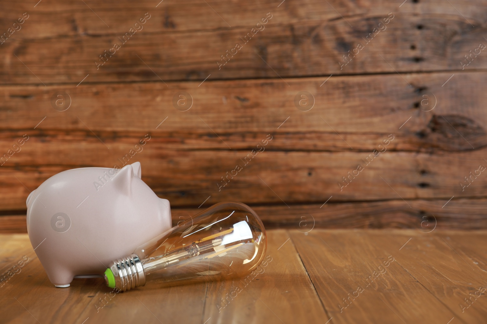 Photo of Piggy bank and light bulb on wooden table, space for text. Energy saving concept