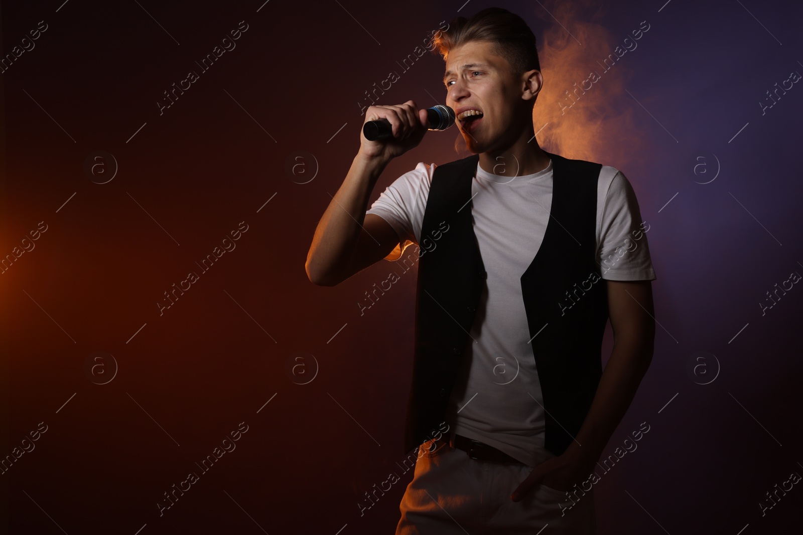 Photo of Talented young man singing on dark background with color light and smoke. Space for text