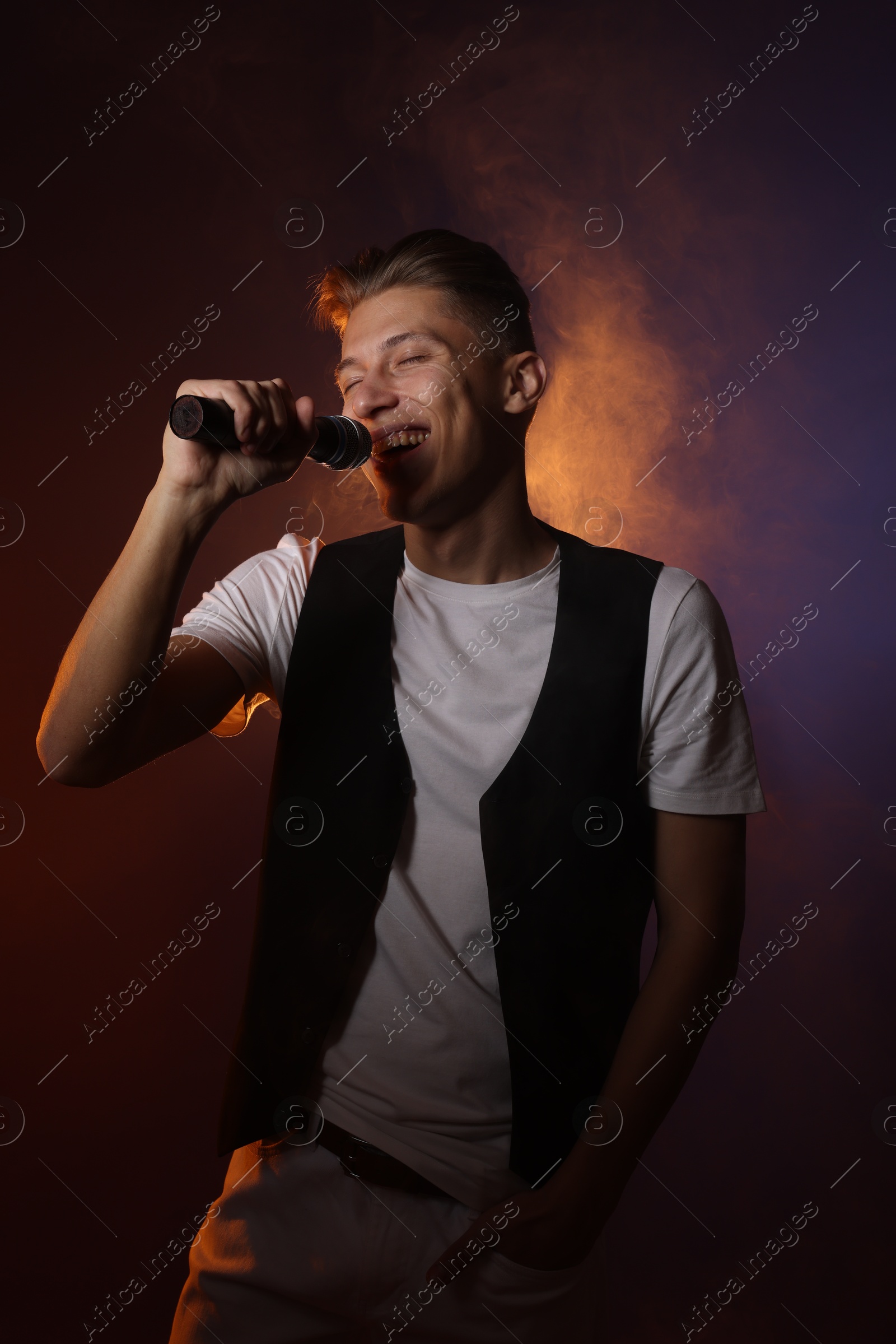 Photo of Talented young man singing on dark background with color light and smoke