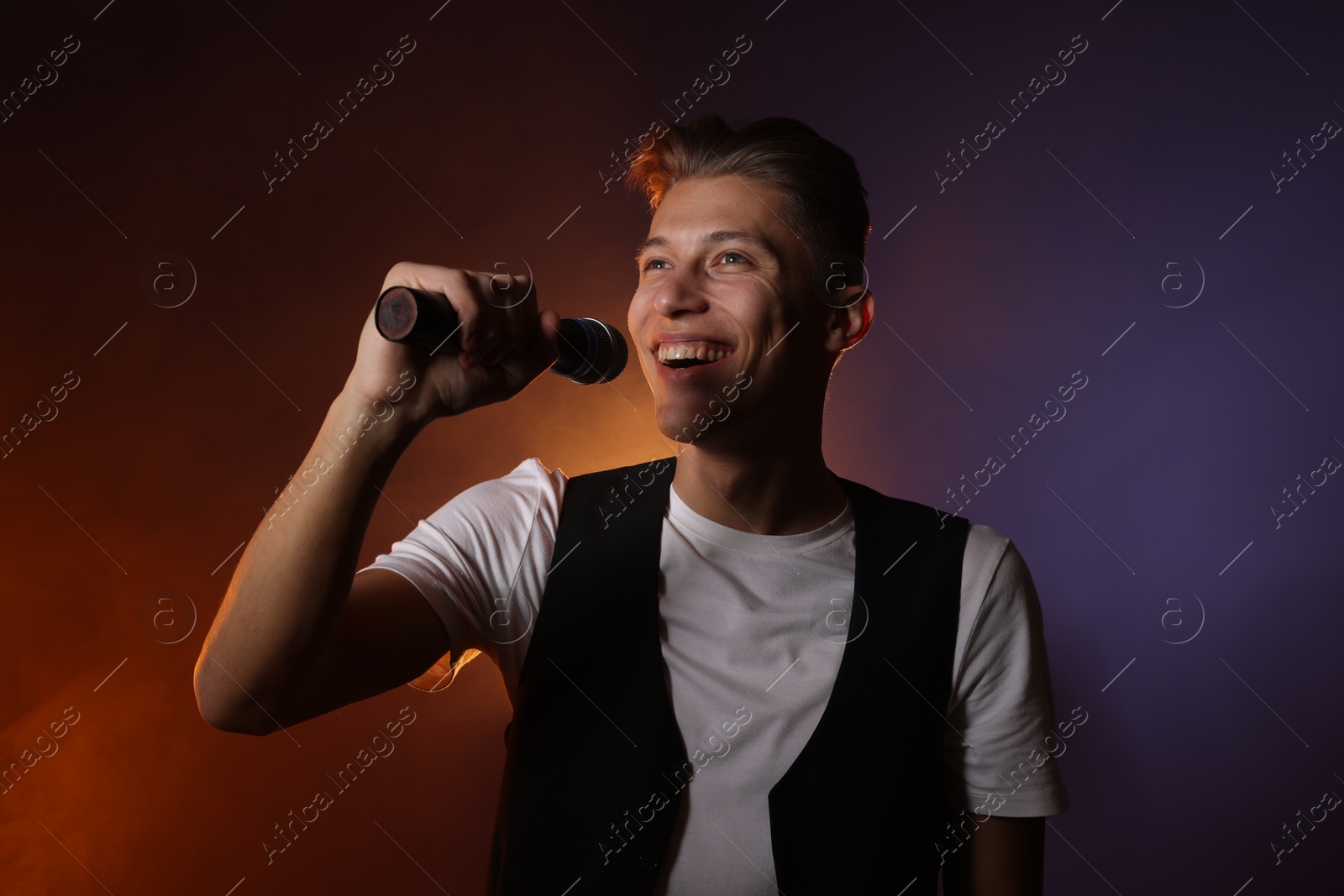 Photo of Talented young man singing on dark background with color light and smoke