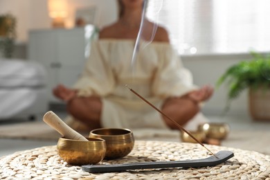 Photo of Young woman practicing yoga on floor indoors, focus on smoldering incense stick and tibetan singing bowls