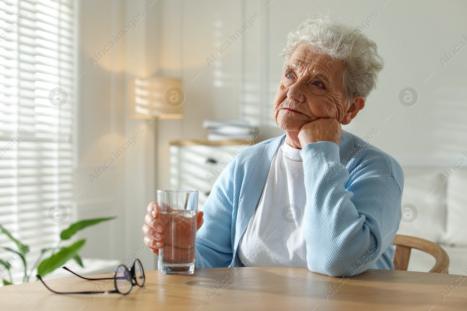 Photo of Thoughtful senior woman feeling lonely at table with glass of water indoors