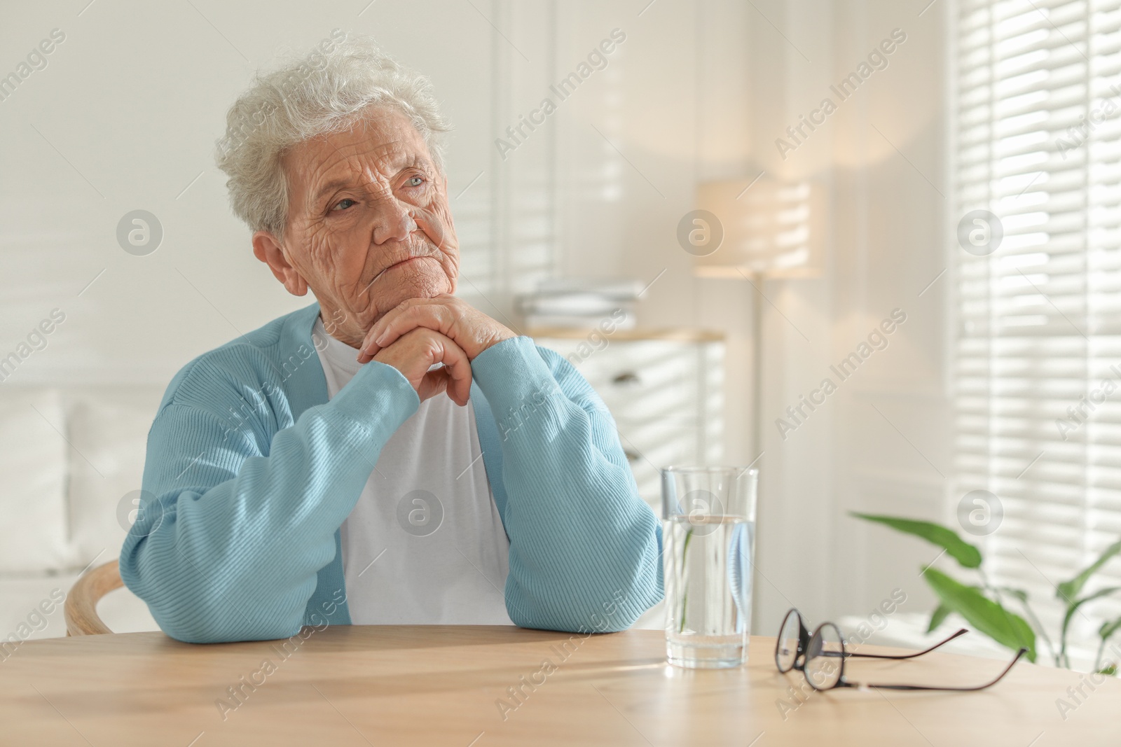 Photo of Thoughtful senior woman feeling lonely at table with glass of water indoors