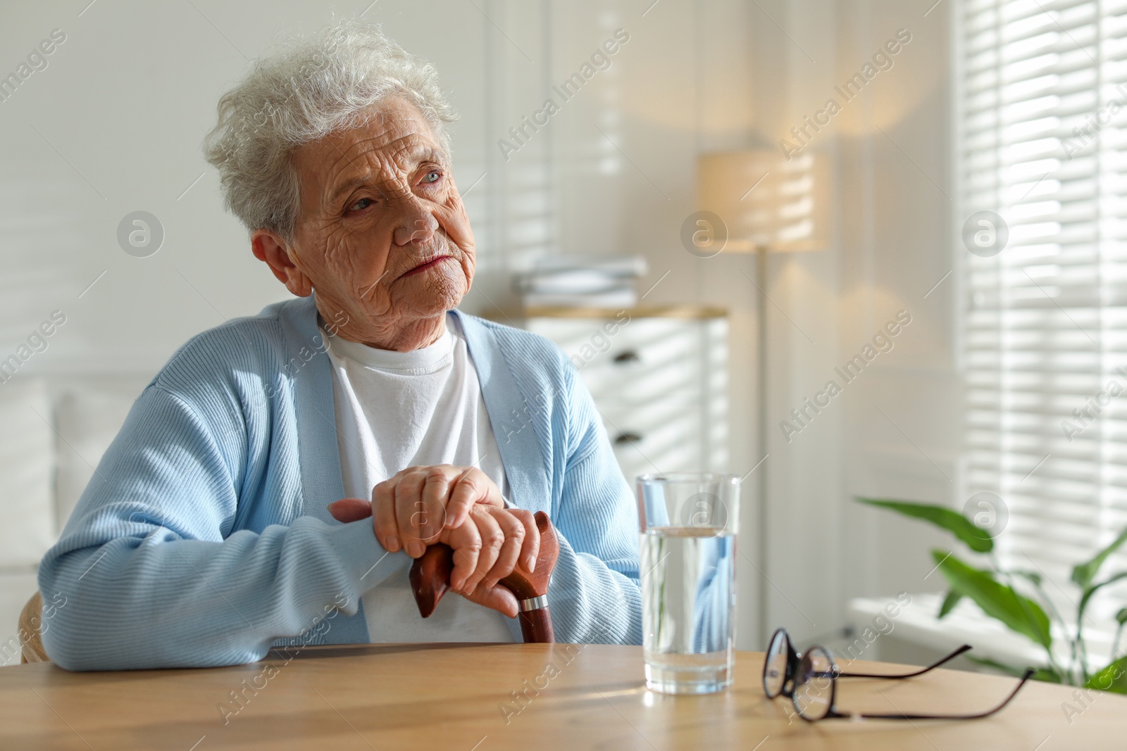 Photo of Thoughtful senior woman feeling lonely at table with glass of water indoors