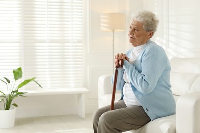 Photo of Lonely senior woman with walking cane sitting on sofa indoors. Space for text