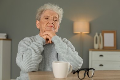 Photo of Sad senior woman feeling lonely at wooden table with cup and glasses indoors