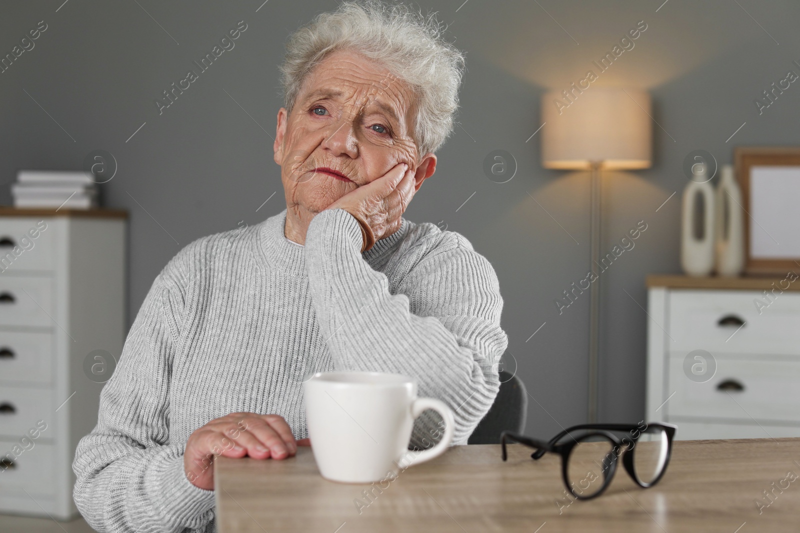 Photo of Sad senior woman feeling lonely at wooden table with cup and glasses indoors