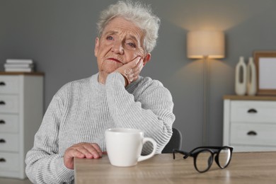 Photo of Sad senior woman feeling lonely at wooden table with cup and glasses indoors