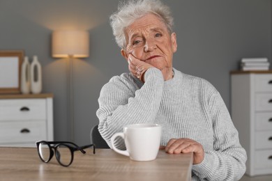 Photo of Sad senior woman feeling lonely at wooden table with cup and glasses indoors