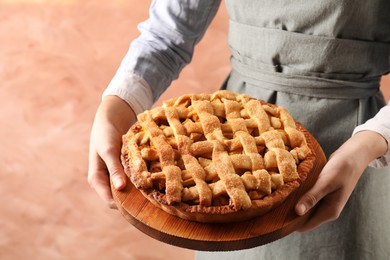 Photo of Woman holding wooden board with tasty homemade apple pie on light coral background, closeup