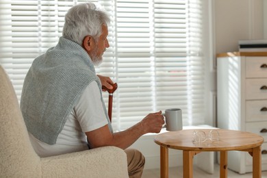 Photo of Lonely senior man with walking cane and cup of drink sitting on sofa at home