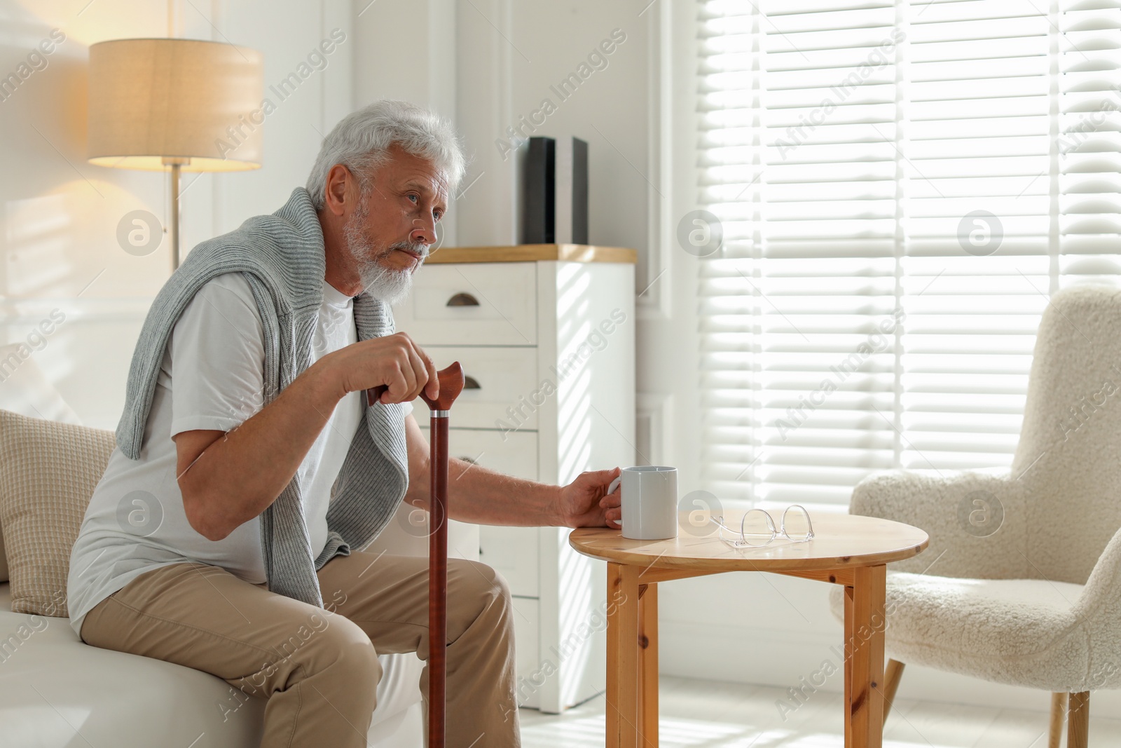 Photo of Lonely senior man with walking cane and cup of drink sitting on sofa at home