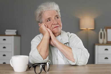 Photo of Sad senior woman feeling lonely at wooden table with cup and glasses indoors