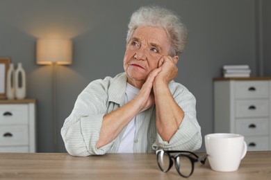 Photo of Sad senior woman feeling lonely at wooden table with cup and glasses indoors