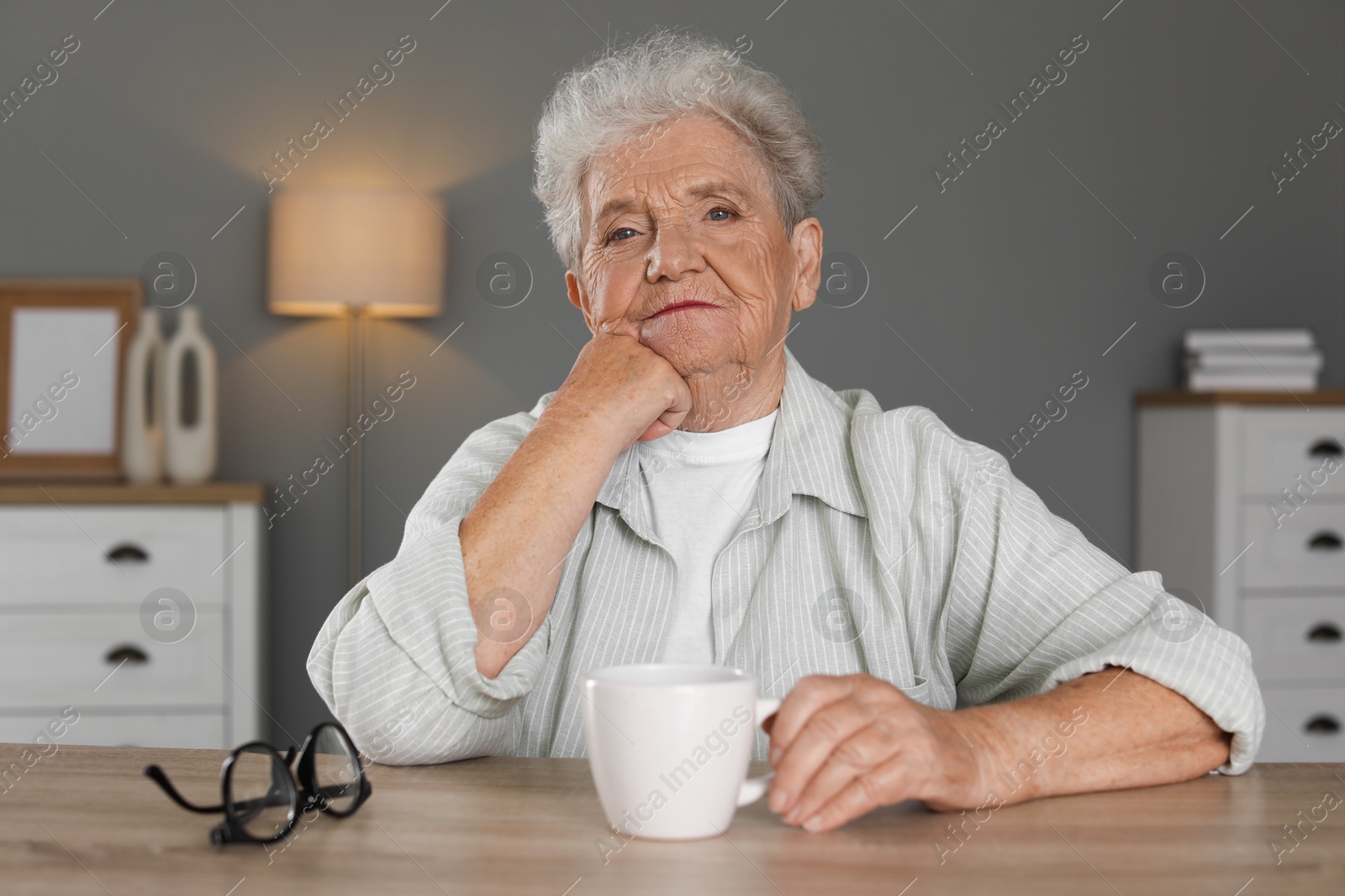 Photo of Sad senior woman feeling lonely at wooden table with cup and glasses indoors