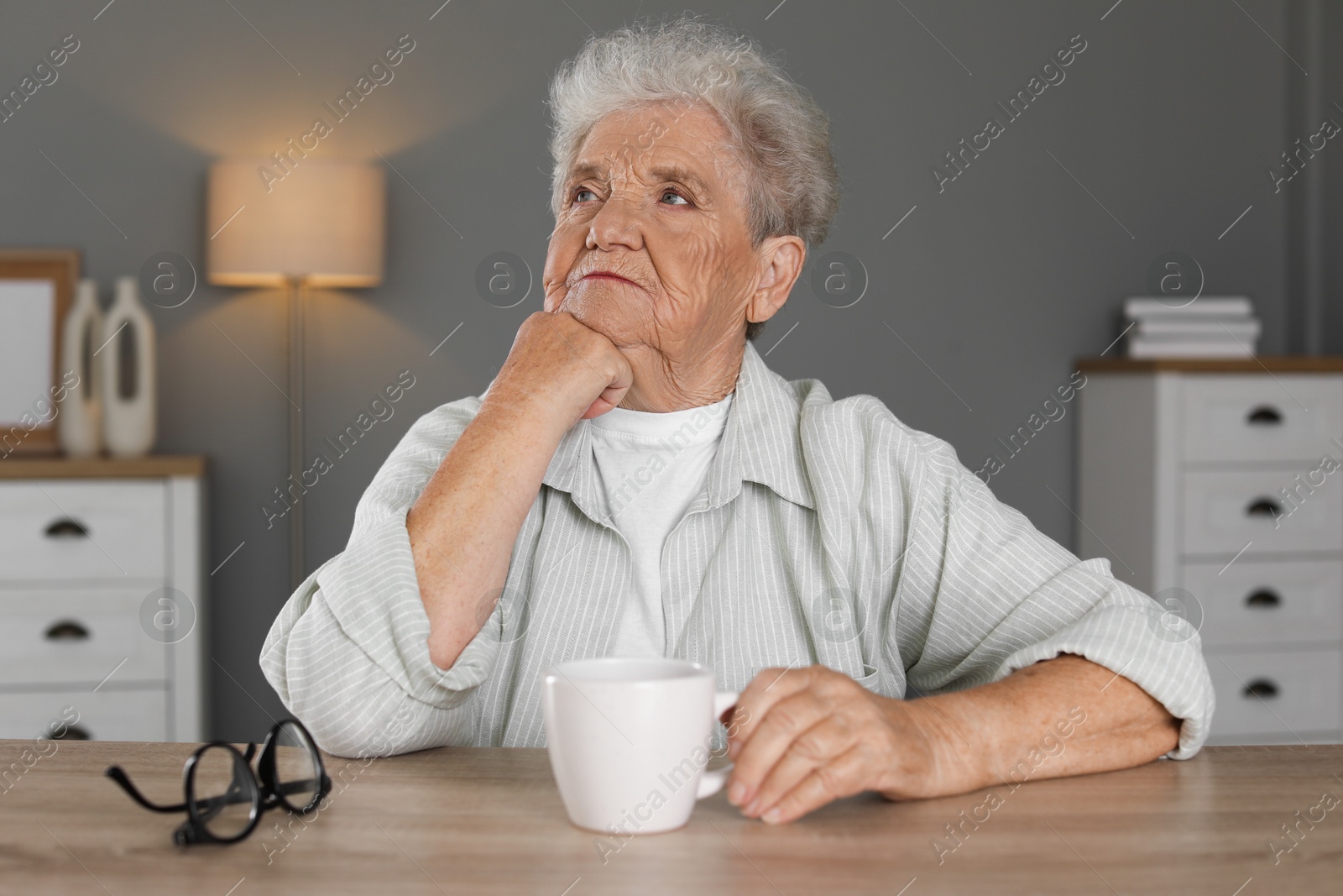 Photo of Sad senior woman feeling lonely at wooden table with cup and glasses indoors