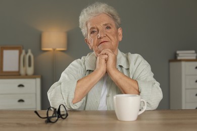 Photo of Sad senior woman feeling lonely at wooden table with cup and glasses indoors