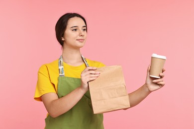 Photo of Girl in apron holding takeaway cup of coffee and paper bag on pink background. Work for teenagers