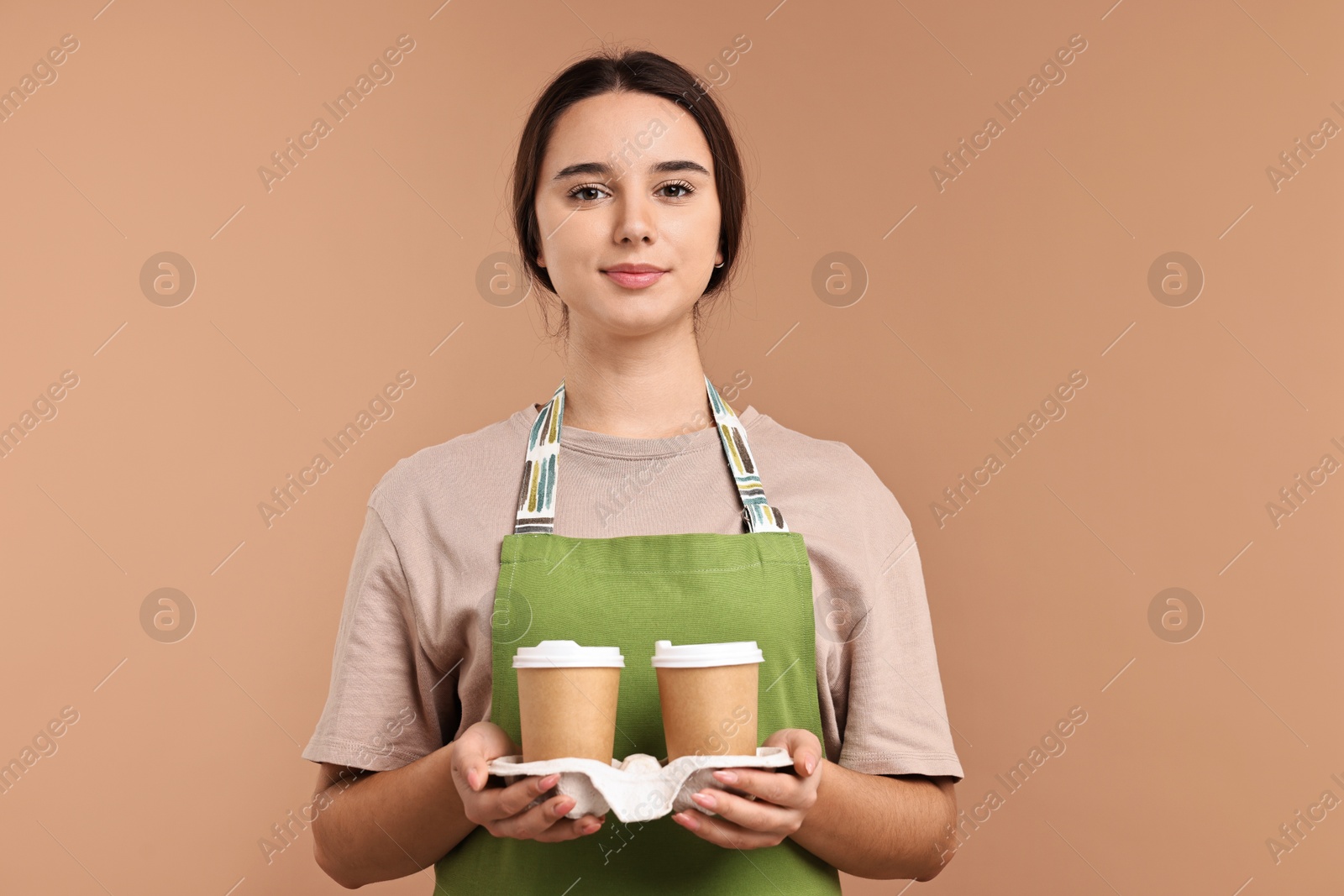 Photo of Girl in apron with takeaway paper cups of coffee on pale brown background. Work for teenagers