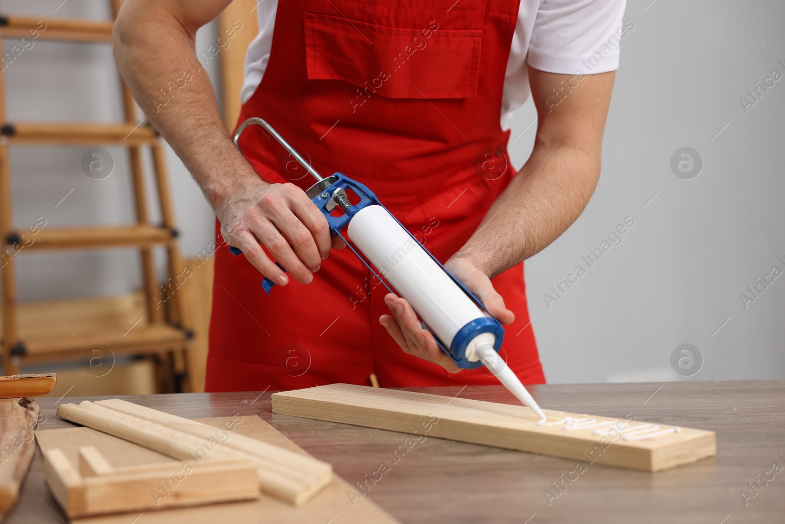 Photo of Worker with caulking gun glueing wooden plank indoors, closeup