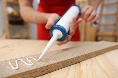 Photo of Man with caulking gun glueing wooden plank indoors, closeup