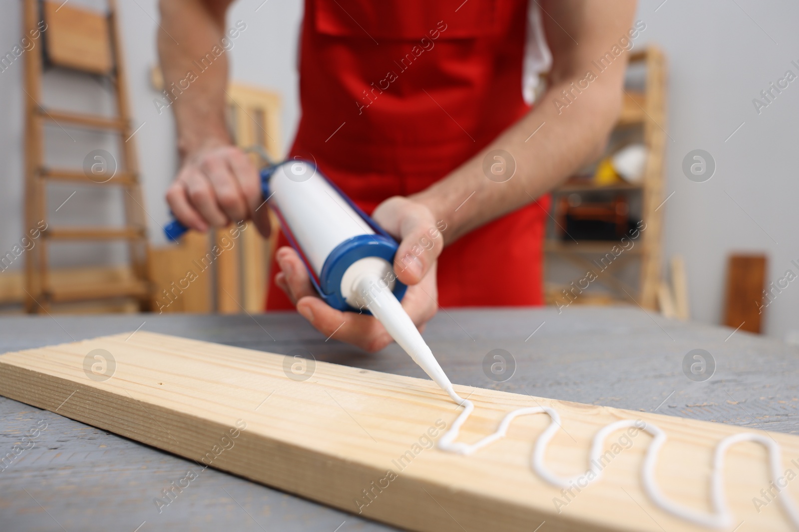 Photo of Worker with caulking gun glueing wooden plank indoors, closeup