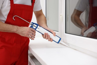 Photo of Worker with caulking gun sealing window indoors, closeup