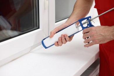 Photo of Worker with caulking gun sealing window indoors, closeup