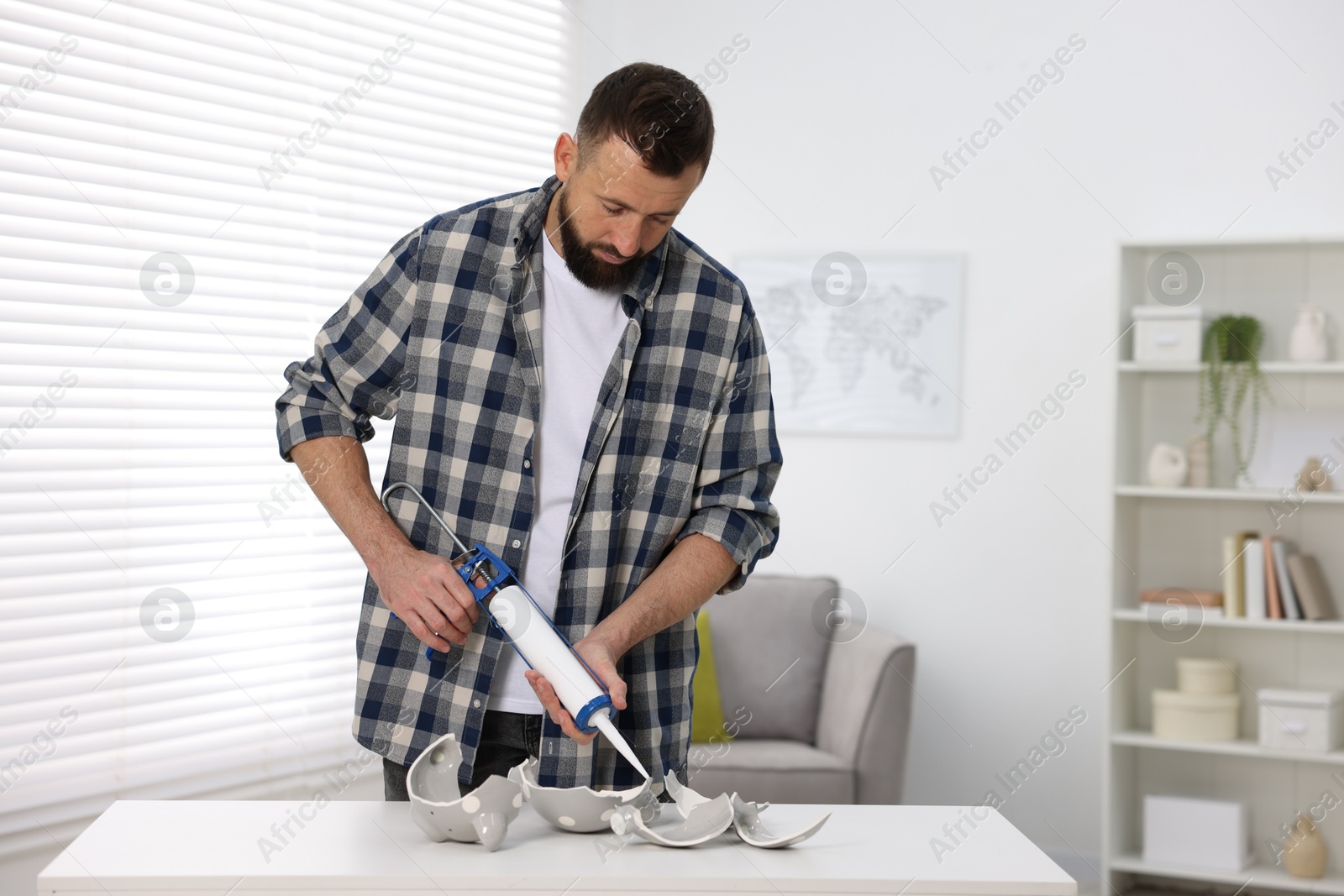 Photo of Man with caulking gun glueing pieces of ceramic piggy bank indoors