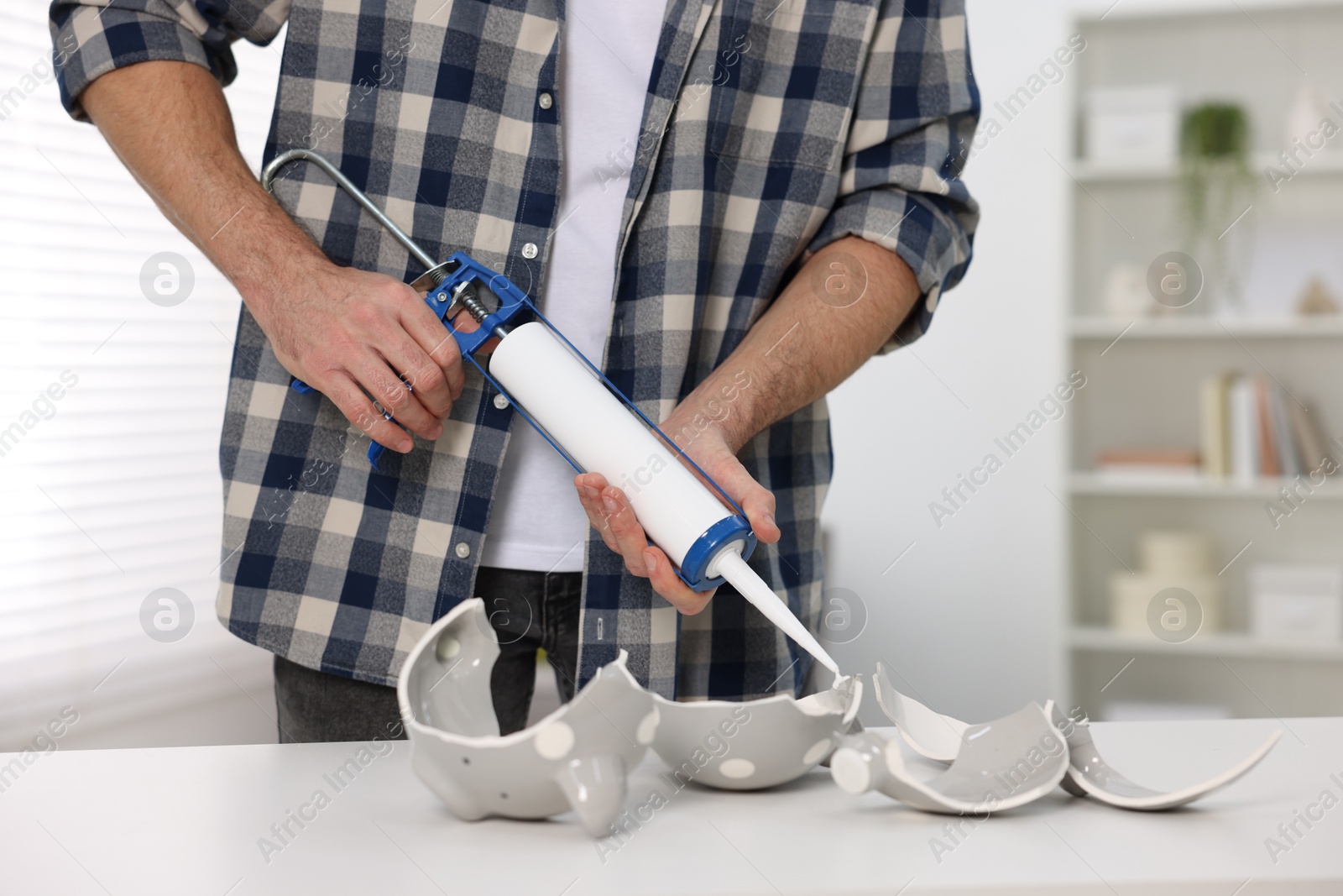 Photo of Man with caulking gun glueing pieces of ceramic piggy bank at white table indoors, closeup