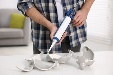 Photo of Man with caulking gun glueing pieces of ceramic piggy bank at white table indoors, closeup