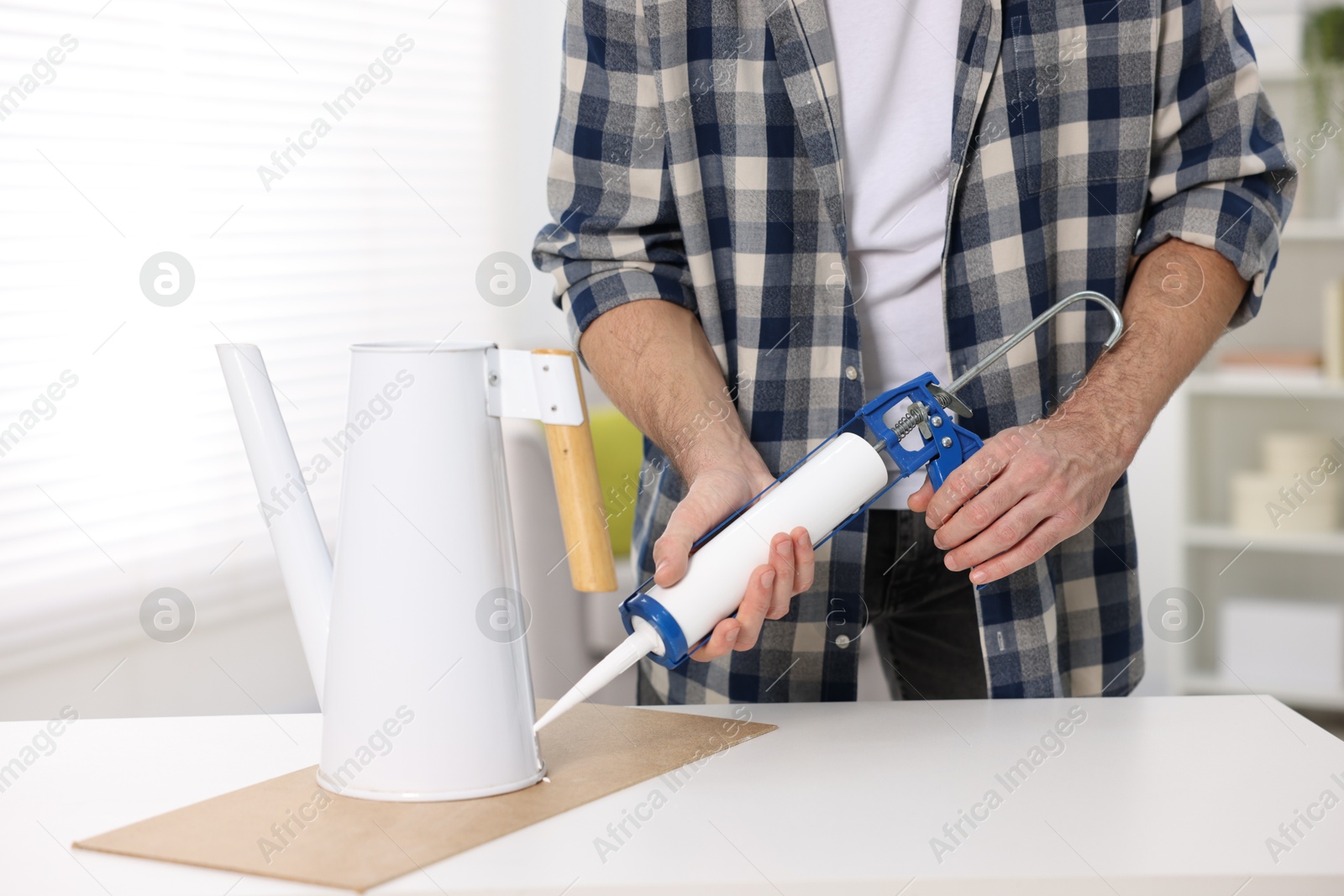 Photo of Man with caulking gun glueing watering can at white table indoors, closeup