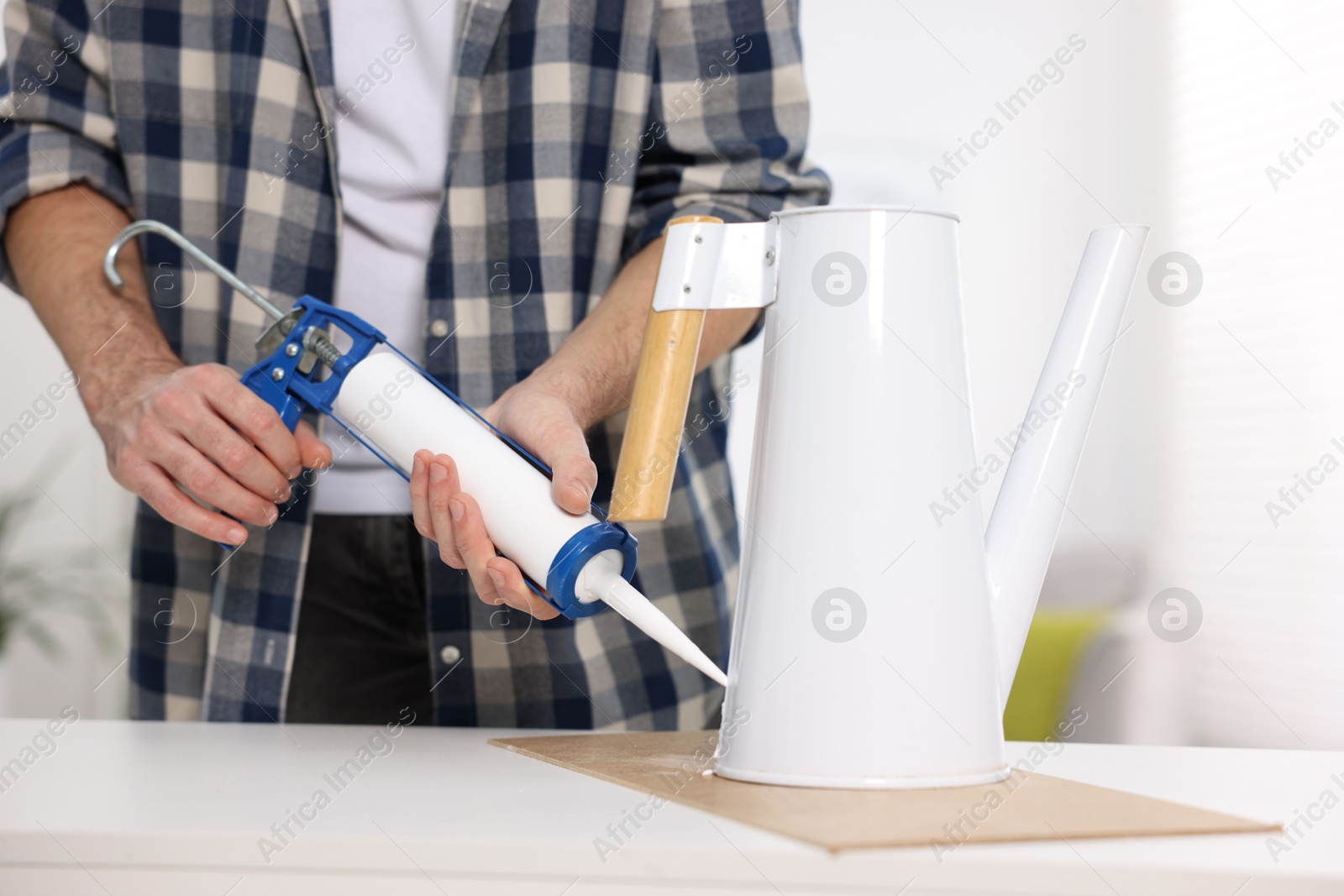 Photo of Man with caulking gun glueing watering can at white table indoors, closeup