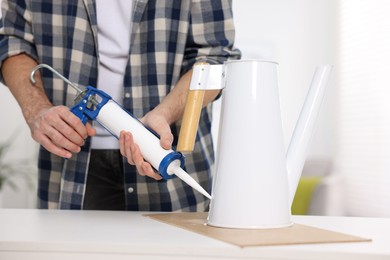 Photo of Man with caulking gun glueing watering can at white table indoors, closeup